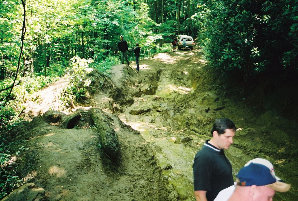 People walking a deeply eroded muddy forest trail with a vehicle parked in the distance