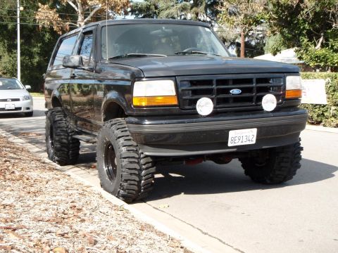 1995 Ford Bronco - This lifted 1995 Ford Bronco is parked curbside on a street with trees in the background.