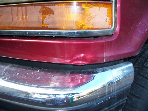 1977 Ford F-150 - Close-up of the front corner of a red 1977 Ford F-150, showing a dented bumper and a dirty headlight.