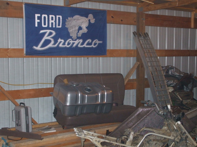 Silver fuel tank on a bench seat in a garage under a blue Ford Bronco flag, with ramps and parts nearby