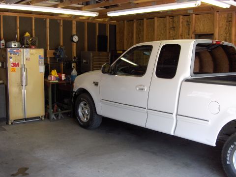 2007 Ford Mustang - White late-1990s Ford F-150 SuperCab pickup parked nose-in inside a stud-framed garage lit by fluorescent shop lights, shown in driver-side…