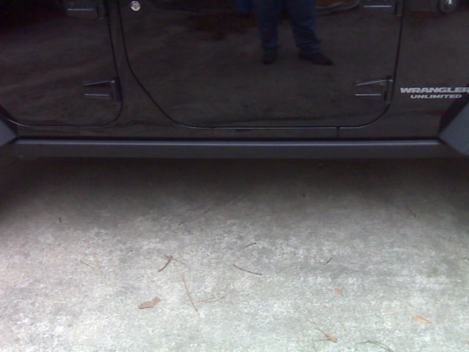 Side of a black Jeep Wrangler Unlimited showing the door and rock rail. The 'Wrangler Unlimited' badge is visible