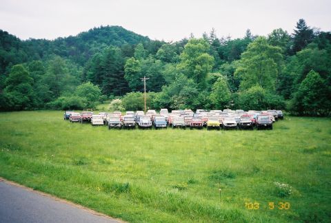 1991 Ford Bronco - A large collection of SUVs and trucks are parked in a field, surrounded by lush green trees and a distant hillside.