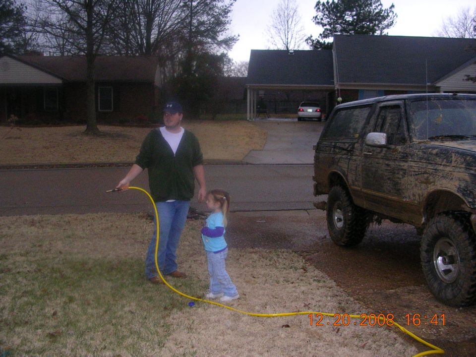 Muddy SUV parked in driveway, man holding yellow hose, girl standing nearby