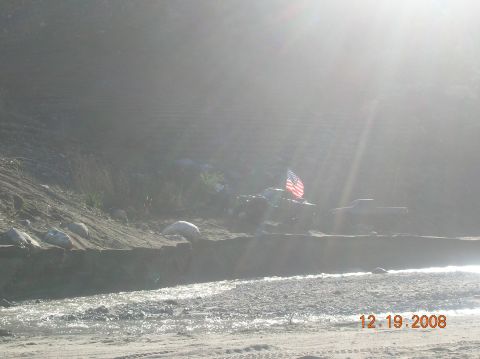 1985 Ford Bronco - An off-road vehicle with an American flag is parked by a river, with another truck visible in the distance.
