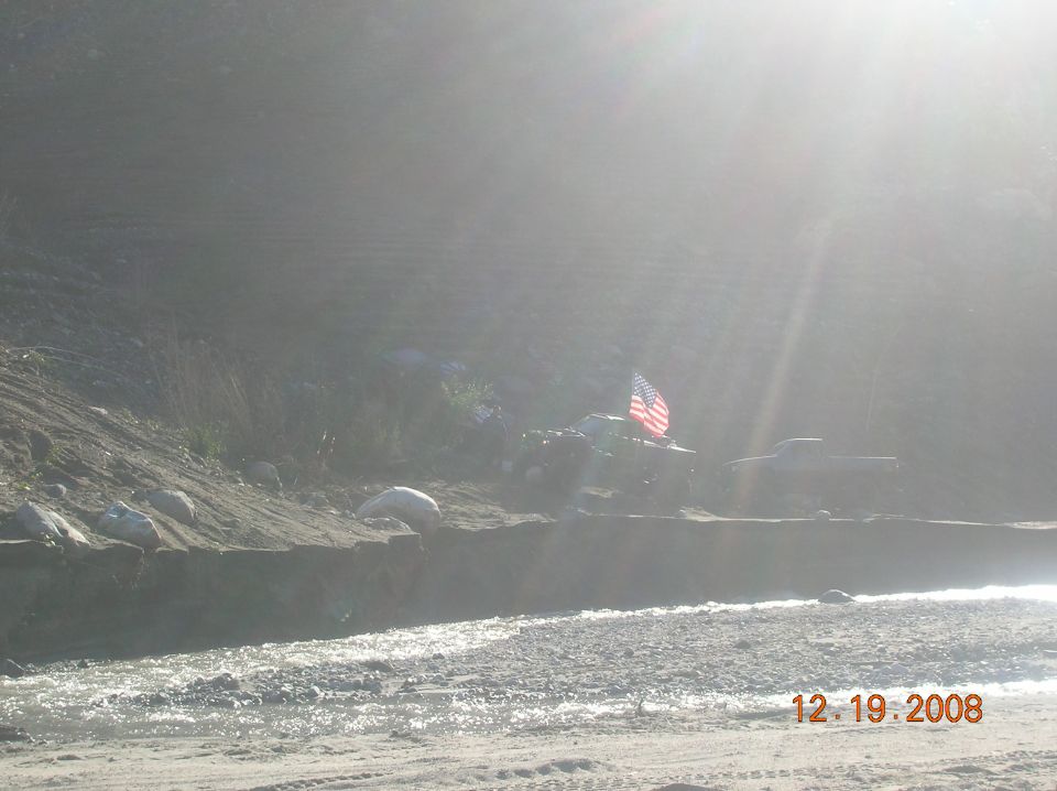 Off-road vehicle with American flag flying, parked near a riverbank with another truck in the background