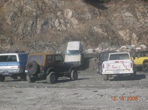 1985 Ford Bronco - Multiple off-road vehicles are parked on a dirt lot, with a rocky hillside behind them.