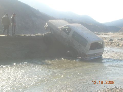 1985 Ford Bronco - A white Ford Bronco is stuck in shallow water, tilted precariously.