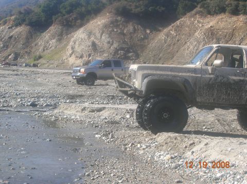1985 Ford Bronco - A muddy off-road vehicle is parked on a rocky riverbed next to another truck.