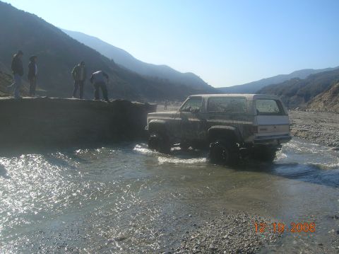 1985 Ford Bronco - A GMC Jimmy navigates a rocky stream bed, with onlookers observing from a dirt embankment.