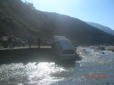 1985 Ford Bronco - Multiple vehicles are submerged in a shallow body of water, with a steep, rocky hillside dominating the background.