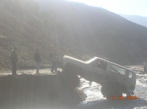 1985 Ford Bronco - A white SUV is stuck in a shallow stream with a muddy bank, while onlookers observe.