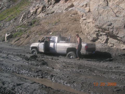 1985 Ford Bronco - A silver pickup truck is deeply mired in mud on a steep, rocky terrain, with a man observing the situation.