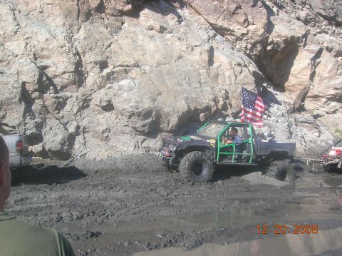1985 Ford Bronco - Off-road vehicle navigates a muddy terrain with a rocky cliff face in the background.