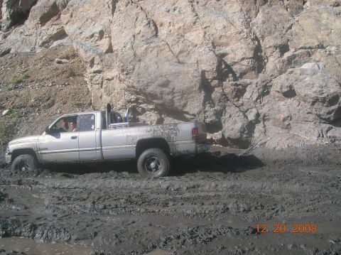 1985 Ford Bronco - A pickup truck is stuck in thick mud on a trail next to a large rock face.
