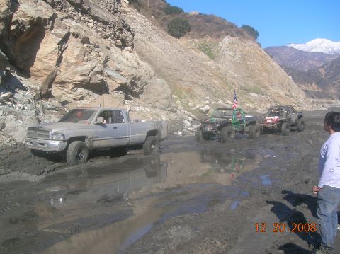 1985 Ford Bronco - Multiple off-road vehicles navigate a muddy riverbed under a clear sky.