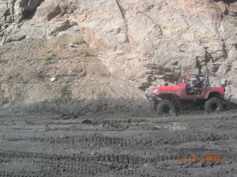 1985 Ford Bronco - A red Jeep Wrangler navigates a challenging muddy terrain with a large rock face in the background.