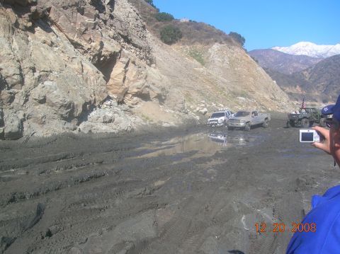 1985 Ford Bronco - Off-road vehicles navigate a muddy terrain with a mountainous backdrop.