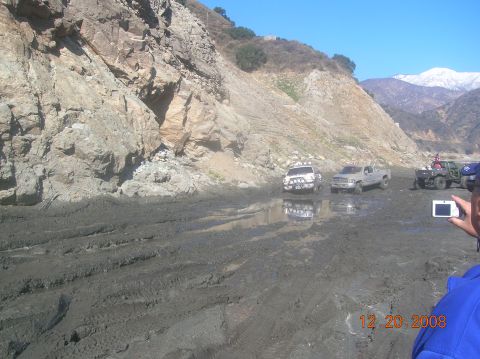 1985 Ford Bronco - Several off-road vehicles navigate a muddy, wet landscape with a rocky cliff face and snow-capped mountains visible.