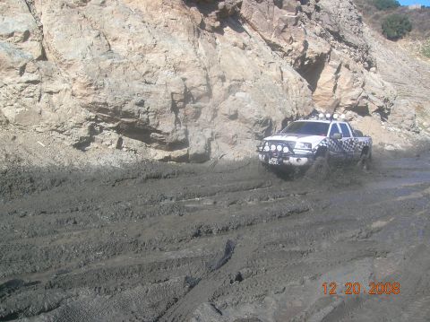 1985 Ford Bronco - A white pickup truck navigates a muddy terrain with a rocky cliff face in the background.
