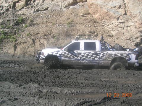 1985 Ford Bronco - A white pickup truck with checkered flag decals is stuck deep in mud, with a rocky hillside in the background. The date 12.20.2008 is visib…