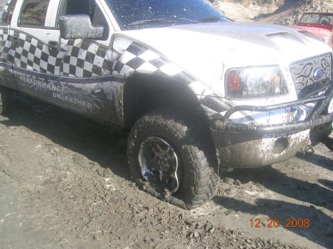1985 Ford Bronco - A muddy Ford F-150 with a flat tire is stuck in the dirt, with a red vehicle visible in the background.