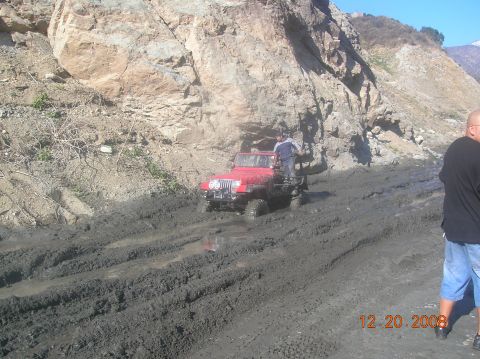 1985 Ford Bronco - A red Jeep Wrangler is stuck in deep mud on a trail, with two people visible in and on the vehicle.