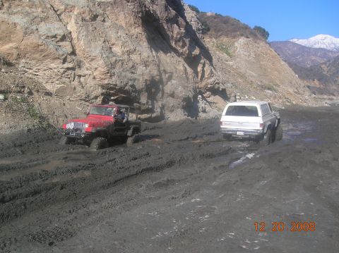 1985 Ford Bronco - Two off-road vehicles are stuck in thick mud on a trail with a large rock face.