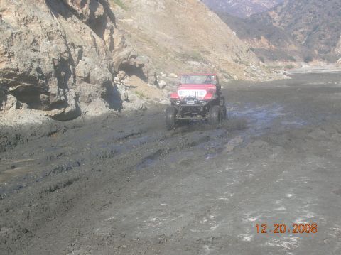 1985 Ford Bronco - A red jeep navigates a muddy terrain with steep hills in the background.