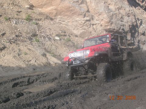1985 Ford Bronco - A red Jeep Wrangler navigates a muddy trail with large tires kicking up dirt.