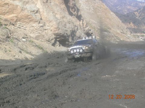 1985 Ford Bronco - A truck navigates a challenging muddy terrain, kicking up spray as it moves forward.