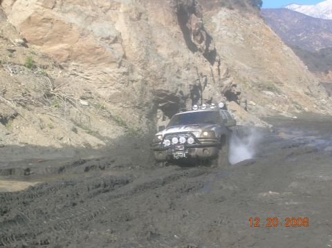 1985 Ford Bronco - A white off-road vehicle navigates a muddy track with a rocky hillside in the background.