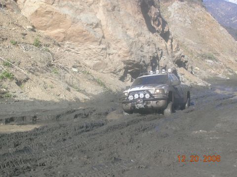 1985 Ford Bronco - A silver pickup truck with multiple auxiliary lights on the front bumper is driving through deep mud and water. The truck is angled towards…