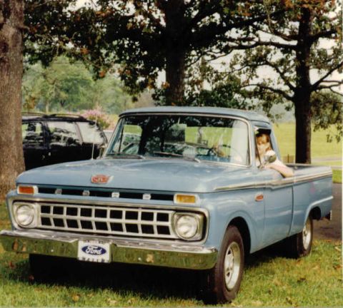 1966 Ford F-100 - A light blue 1960s Ford pickup truck is parked on a grassy area under trees. A young person is leaning out of the driver's side window.