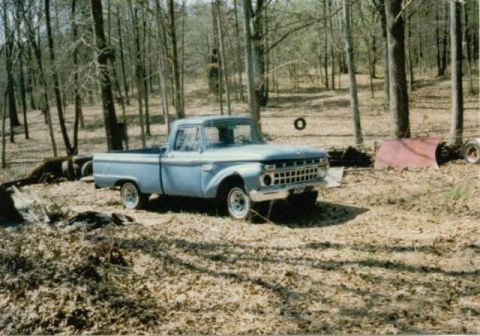 1966 Ford F-100 - A light blue 1960s era Ford pickup truck is parked in a wooded area with fallen leaves on the ground. A red tarp and a tire are visible in…
