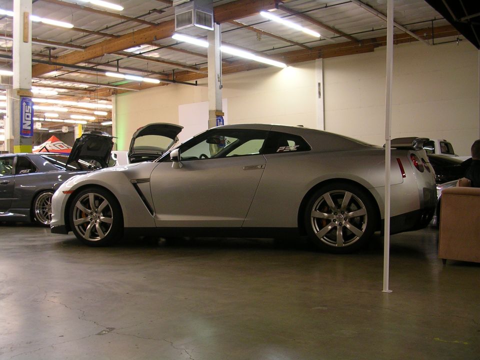 Silver Nissan GT-R parked indoors with hood open, next to a dark gray sedan.