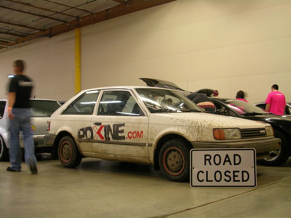 White Mazda hatchback with "BOXINE.COM" text and a "ROAD CLOSED" sign in front.