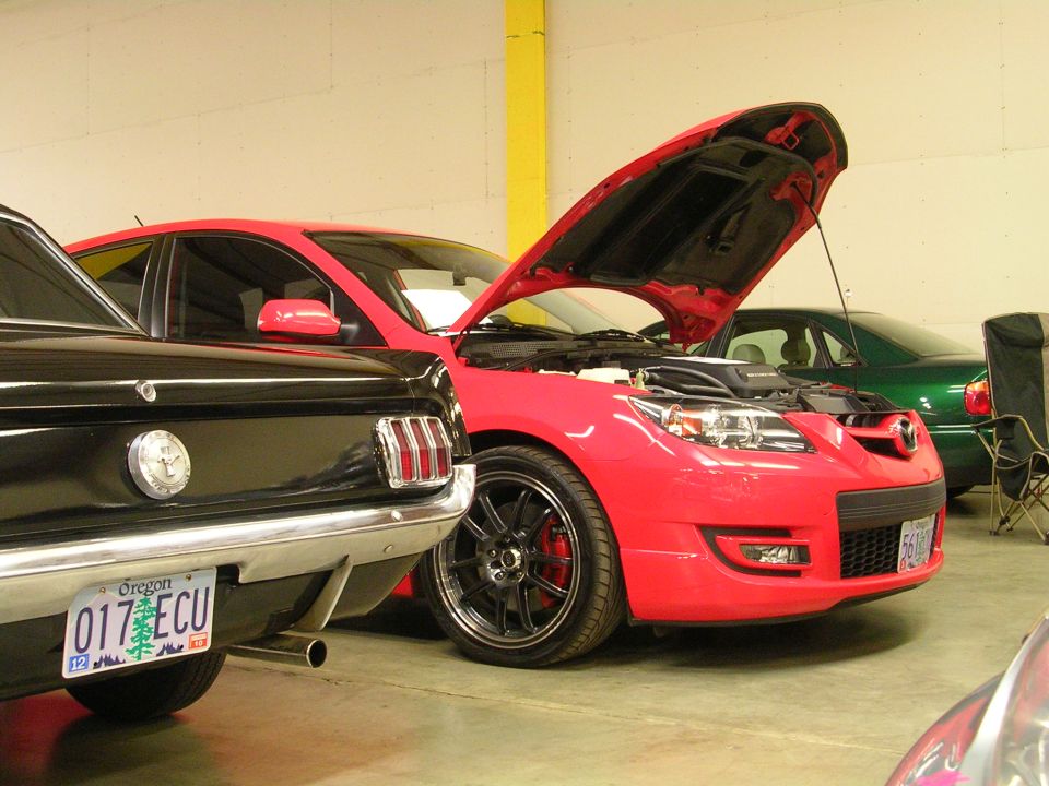Red Mazda 3 with hood open next to a black Ford Mustang, green sedan in background.