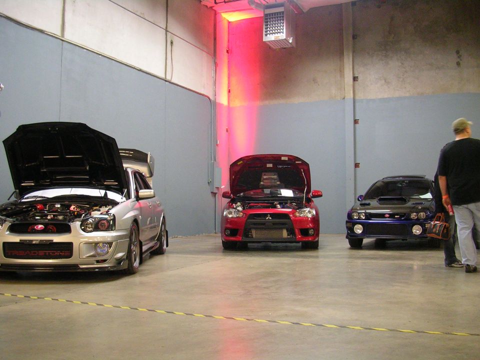 Silver Subaru WRX STI, red Mitsubishi Lancer Evolution, and dark blue Subaru WRX STI with hoods open.