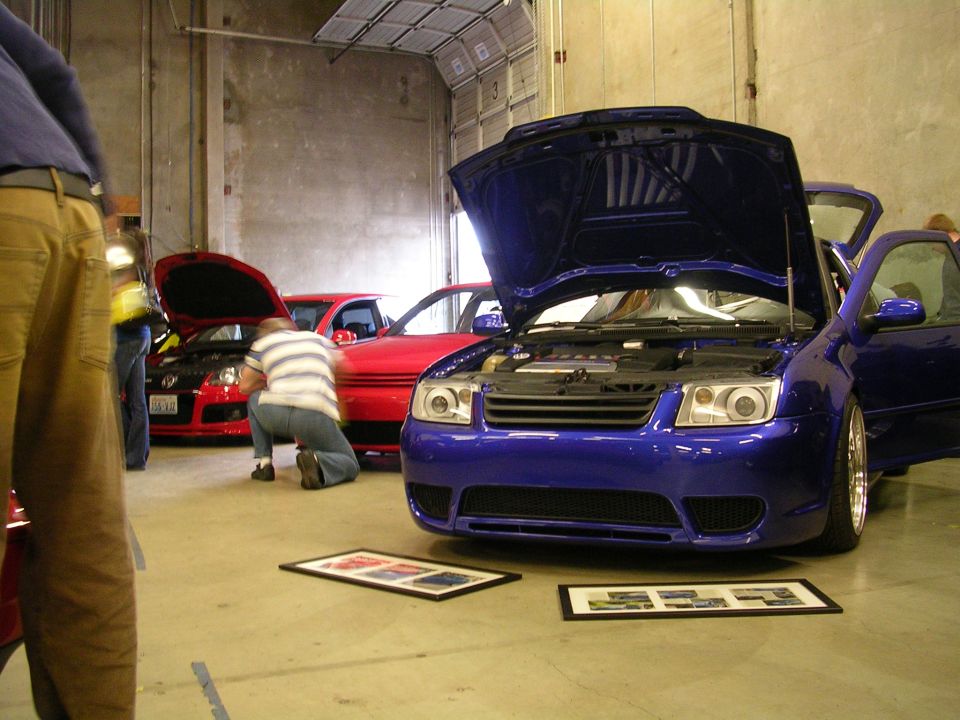 Blue Volkswagen Jetta with hood and door open, red Jetta behind, person kneeling.