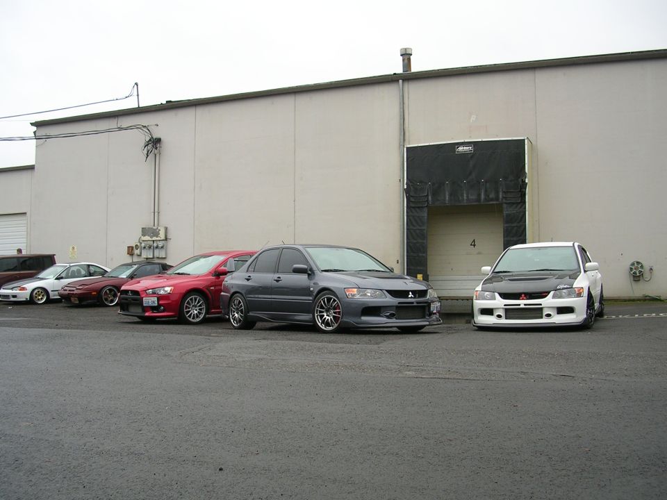 White and grey Mitsubishi Lancer Evolutions parked in front of a loading dock with other cars.