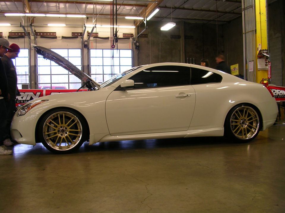 White two-door coupe with gold wheels, hood open, in a garage.