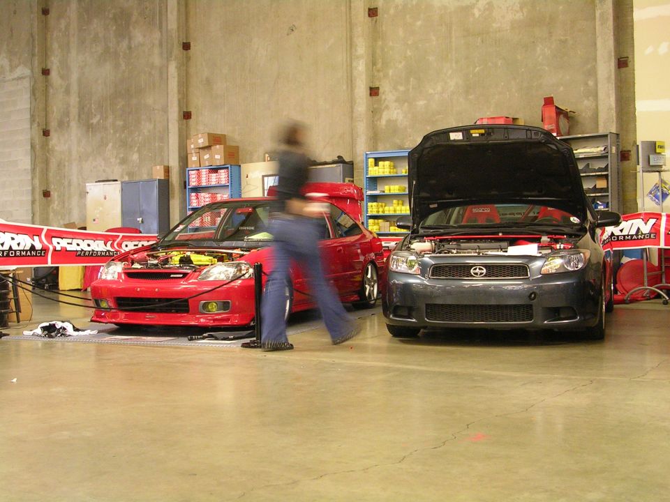 Blurred person in front of red car with open hood; dark gray car with open hood.