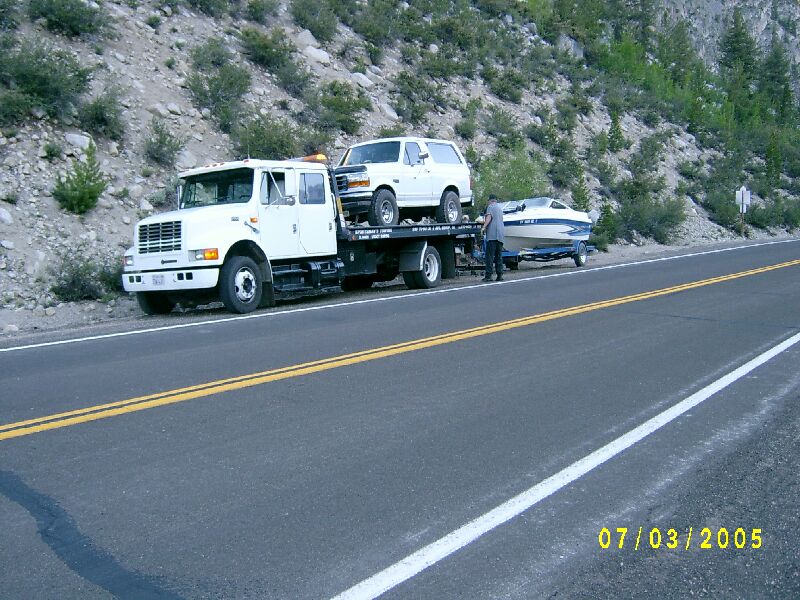 White Ford Bronco on a flatbed tow truck towing a boat trailer along a mountain roadside