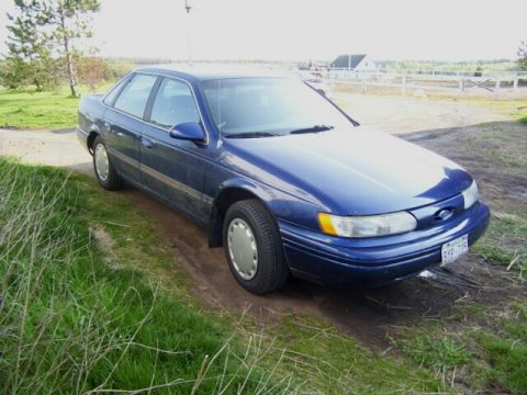 1995 Ford Taurus - A blue 1995 Ford Taurus sedan is parked on a dirt path next to a grassy area.