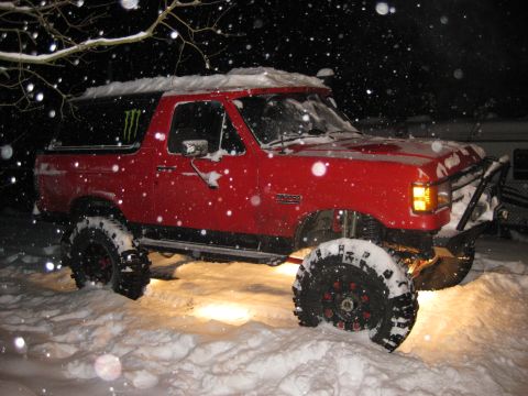 1979 Ford Bronco - A lifted red Ford Bronco is parked in the snow at night, with snow falling. The vehicle has large off-road tires and a Monster Energy stick…