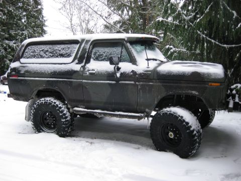 1979 Ford Bronco - A dark-colored, lifted Ford Bronco covered in snow sits parked in a snowy, wooded area. Icicles hang from the front fender and the vehicle…