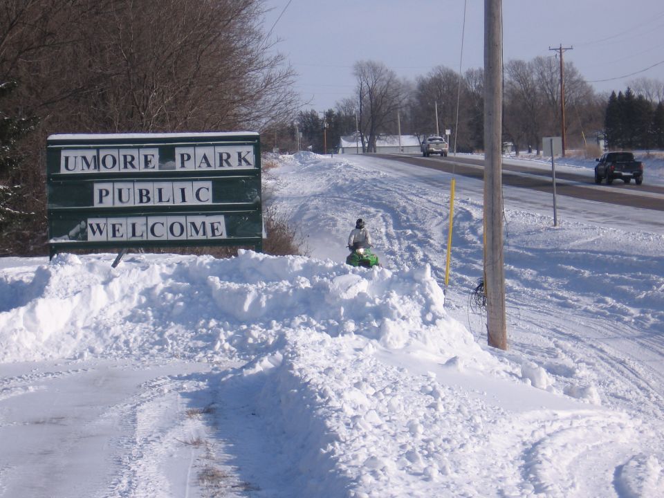 Green snowmobile riding a snowbank trail past a UMORE Park Public Welcome sign