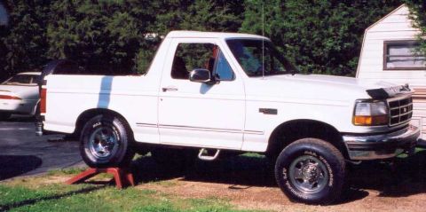 1993 Ford Bronco - A white Ford Bronco is parked in a yard with a trailer in the background. The vehicle is raised on a jack stand.