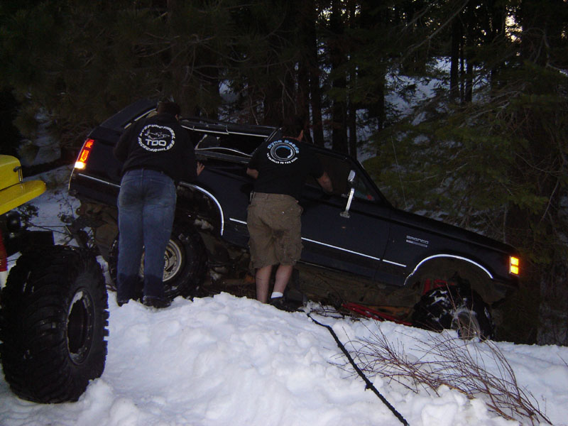 Two people pushing a black lifted truck on a snowy slope at dusk